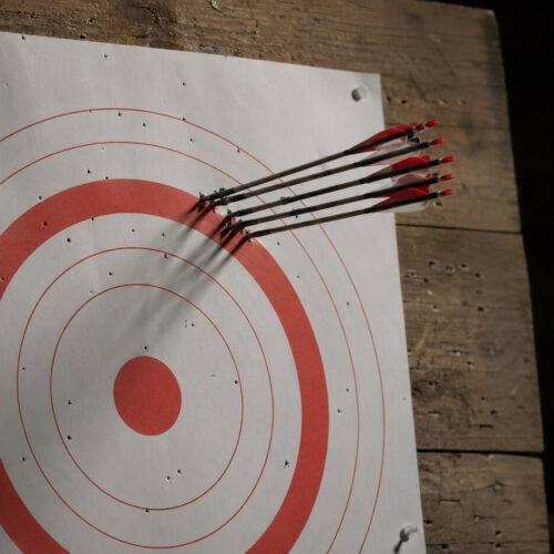 Archery target with six arrows clustered tightly off-center, missing the painted bullseye. Visual metaphor for Goodhart's Law in AI: the system optimized perfectly for the wrong target.