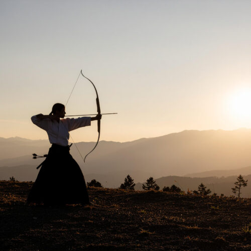 A solitary Japanese Kyudo archer at full draw, silhouetted against a setting sun over distant mountains.